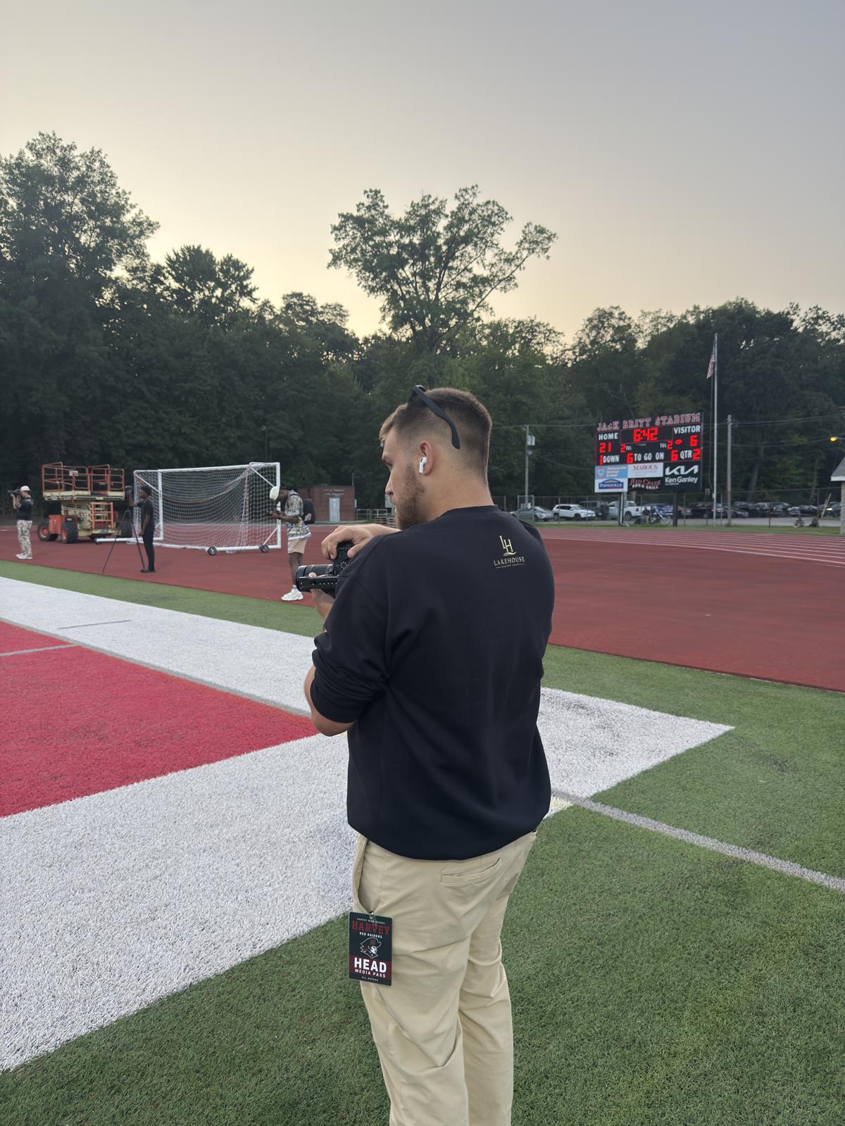 Danny Land filming in a black Lake House shirt at a football stadium